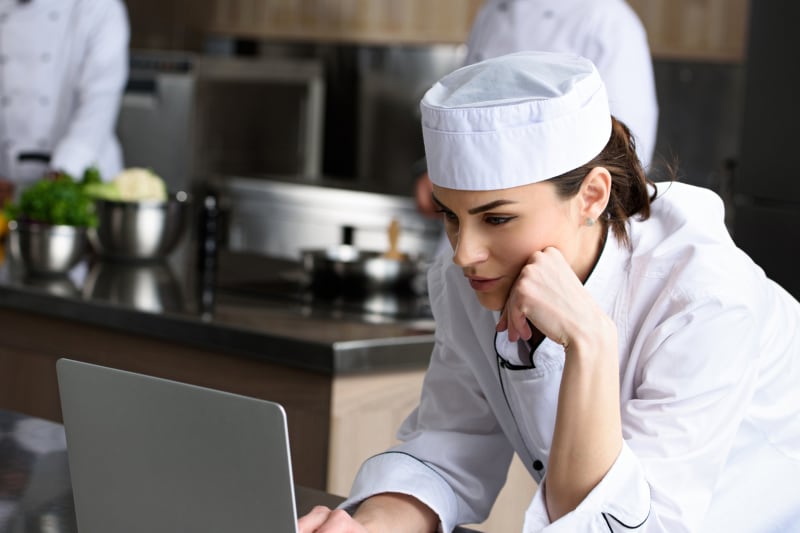 Kucharka w białym stroju sprawdza coś na laptopie w kuchni restauracji
